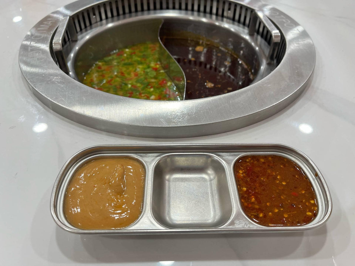 Divided hot-pot broth beside stainless sauce ware with two filled outer wells and an empty center compartment.