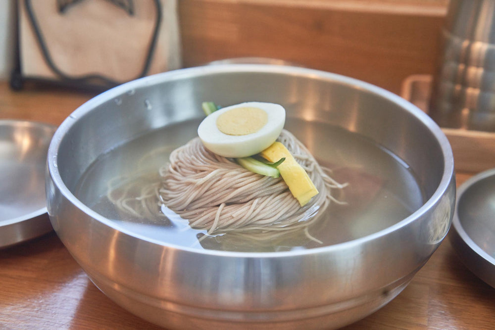 Cold noodles, broth, egg and vegetables fill a broad silver stainless bowl on a restaurant table.