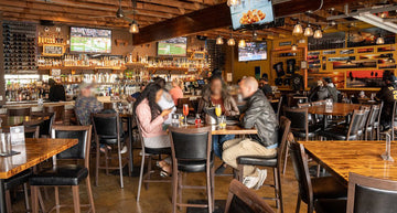 Black Upholstered Restaurant Barstool with Wood-Grain Metal Frame in a restaurant setting; other furniture is illustrative