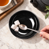 Black shallow dish holding wrapped sugar and a teaspoon during tea service