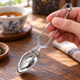 HST013 closed stainless steel tea infuser tongs held for inspection; cup and tabletop props illustrative.