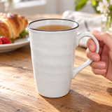 Tall white ceramic mug with dark rim in a table setting. Contextual accessories are not included.