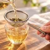 KB8722 fine-mesh tea strainer straining tea into a cup; cups, pitcher and tabletop props illustrative.