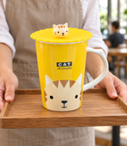 Yellow cat mug with scalloped lid and cat knob in a carrying setting. Contextual accessories are not included.