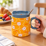 Orange bear mug with blue-gray lid and spoon in a breakfast setting. Contextual accessories are not included.