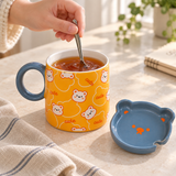 Orange bear mug with blue-gray lid and spoon in a stirring setting. Contextual accessories are not included.