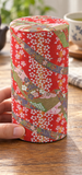 Red ribbon-and-blossom tea container in a table setting. Contextual accessories are not included.