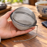 TS85-R closed stainless mesh tea ball supported in a hand, with its chain and hook; cup and table props illustrative.