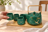 JHS8-GR green and gold tea set with one of four cups held above its plain bamboo tray; surrounding accessories are contextual.