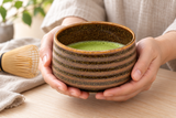 K537-BR brown-banded matcha bowl held in both hands with matcha; whisk and table accessories are contextual.