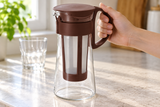 HARIO MCPN-7CBR 600 ml cold-brew pitcher held by its brown handle with the internal filter visible; glass and accessories are contextual.