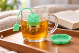 Green owl infuser steeping in a glass mug with matching tray alongside