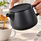 Black coffee dripper pot pouring coffee with a finger securing its flat lid; cups and other props not included.