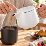 White coffee dripper pot pouring coffee with a finger securing its flat lid; cups and other props not included.