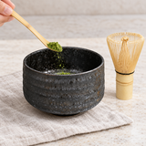 Matcha powder being added to the dark charcoal bowl with bamboo whisk alongside