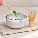 Matcha powder being added to the white blue-rim bowl with bamboo whisk alongside