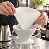 White HARIO V60 size 02 paper filter being opened above a dripper; brewing accessories shown for context, not included.