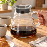 HARIO size 03 clear glass coffee server held by its glass handle at a coffee table; cups and other props not included.