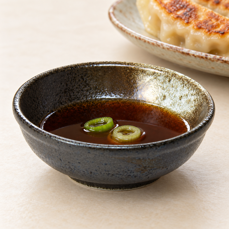 The SKU31 black-and-brown porcelain sauce bowl holding dark sauce with scallion, with dumplings on a separate plate behind it.