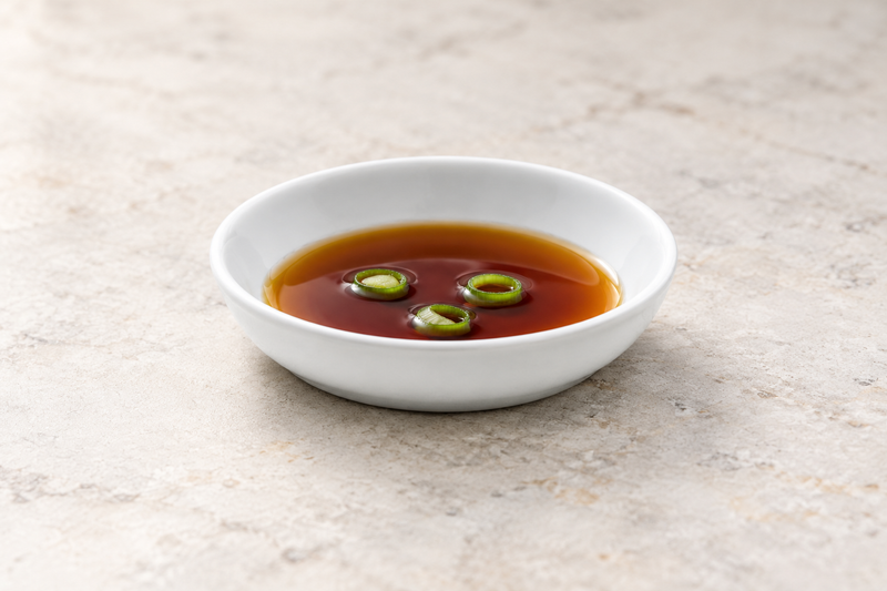 White A1887 sauce dish holding a brown dipping sauce with green garnish on a light stone tabletop.