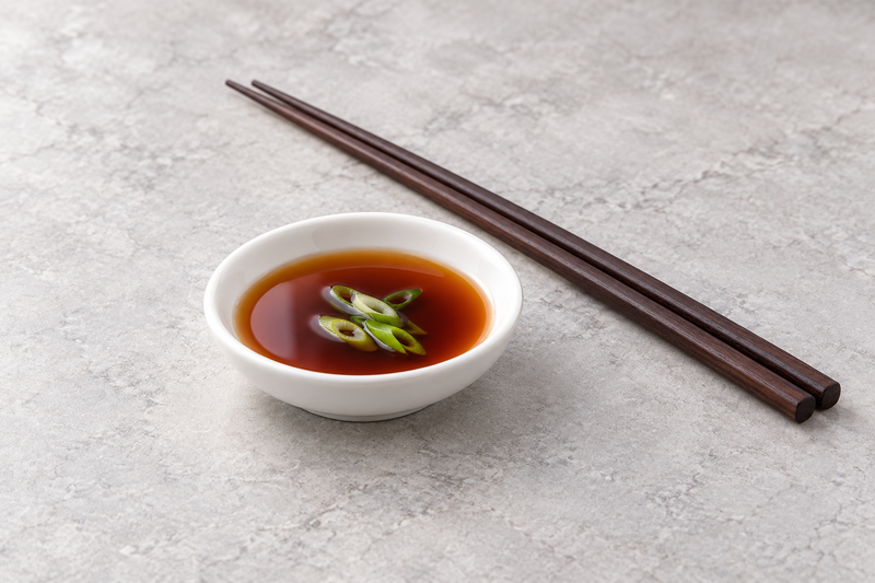 A5544 holding dark sauce with sliced green onion, with brown chopsticks beside it on a pale textured surface.