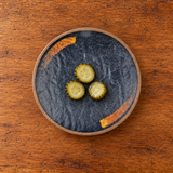 7-inch dark melamine round plate with a small pickle serving on a wooden table
