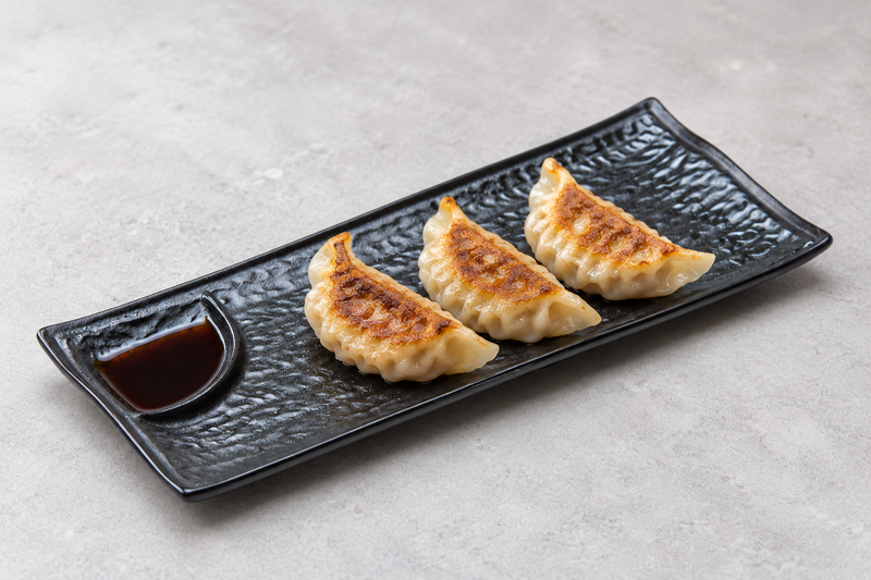 Black divided rectangular plate holding three dumplings beside a sauce-filled end well.