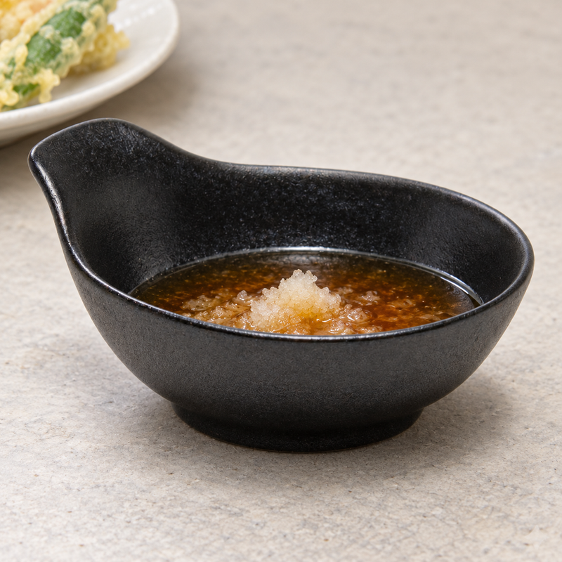 Matte-black handled sauce bowl with brown dipping sauce and pale grated garnish, with a plate of tempura nearby.