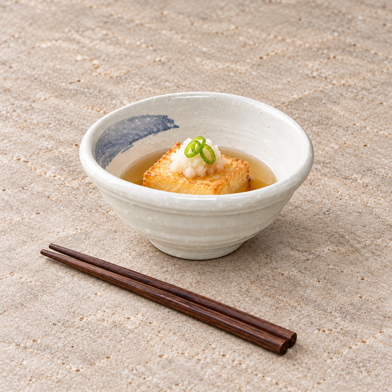Off-white and blue-gray mini bowl holding a small sauced serving with green garnish beside brown chopsticks.