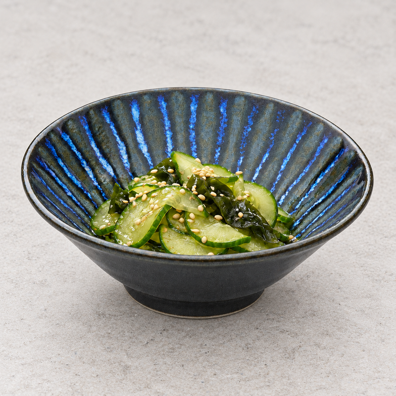 Dark blue radial-pattern shallow bowl holding sliced cucumber and wakame salad sprinkled with sesame seeds.