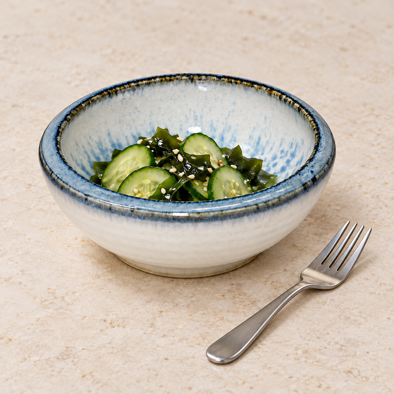 Pale blue and white ceramic bowl holding sliced cucumber and greens, with a fork on a light tabletop.