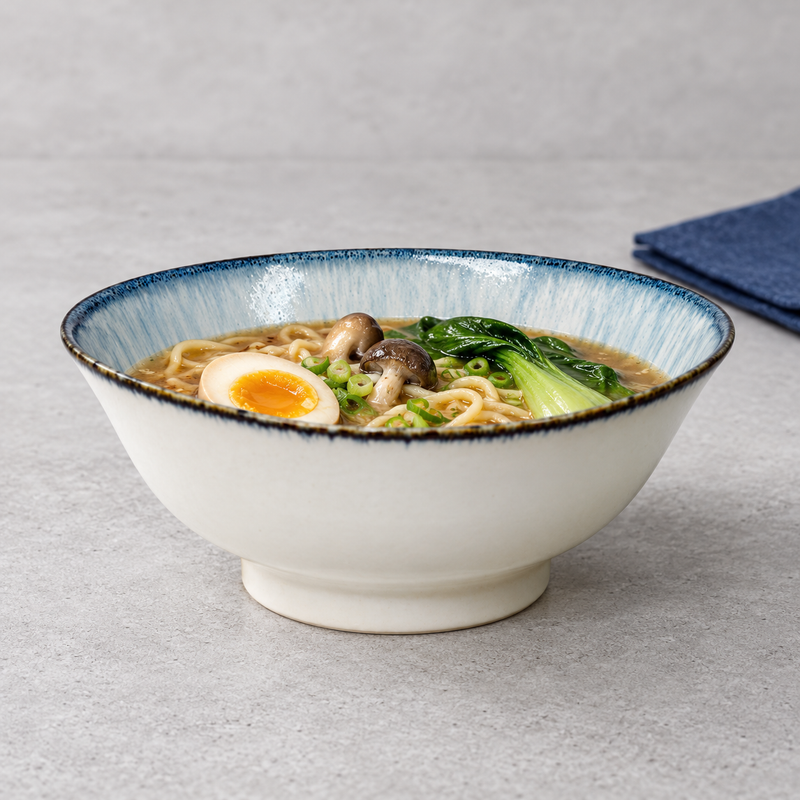Pale blue and white noodle bowl with noodles, green vegetables, mushrooms and a halved egg on a light tabletop.