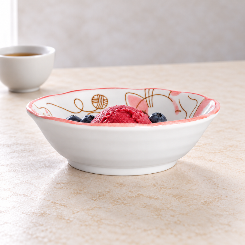 Pink cat-pattern ceramic bowl holding one bright red frozen-dessert scoop and blueberries, with a separate pale cup of amber liquid behind it on the left.