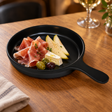 Black round handled melamine serving plate presenting a cold appetizer of prosciutto, pear and cheese