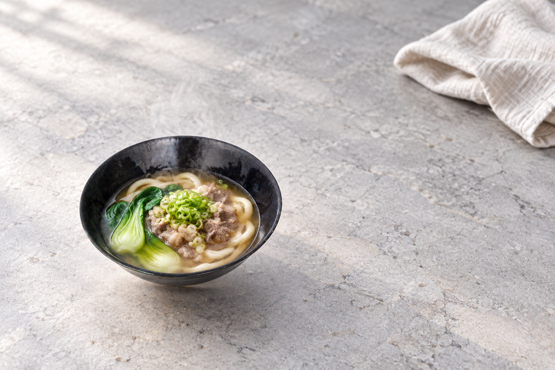 Blue-black bowl holding a noodle soup with green vegetables, meat and chopped scallions in a full tabletop setting.