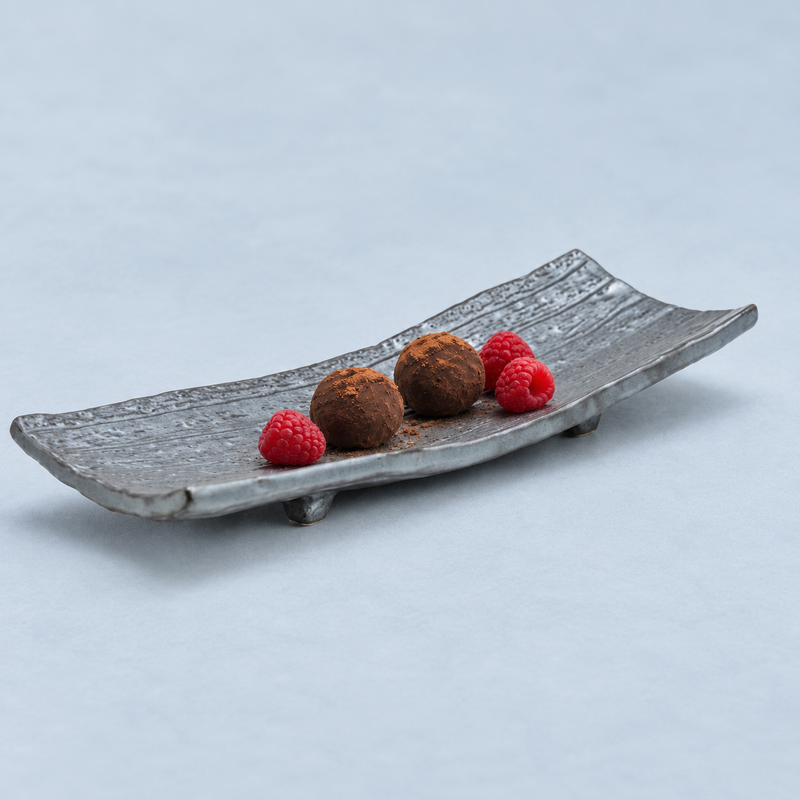 Black elongated porcelain plate holding two cocoa-dusted round sweets and three raspberries.