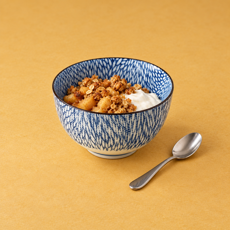 Navy-patterned ceramic bowl with golden fruit chunks, oat-like crumble and a white creamy portion on the right; a metal spoon lies outside lower-right on a mustard background.