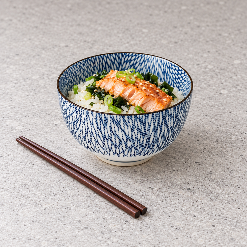 Patterned ceramic bowl with an orange-pink fish fillet over white rice and dark leaves, scallion rings and seeds; dark chopsticks lie diagonally in the lower-left foreground.