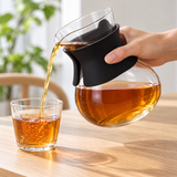 Black-sleeved TDC-50-B glass tea decanter pouring amber tea into a separate drinking glass