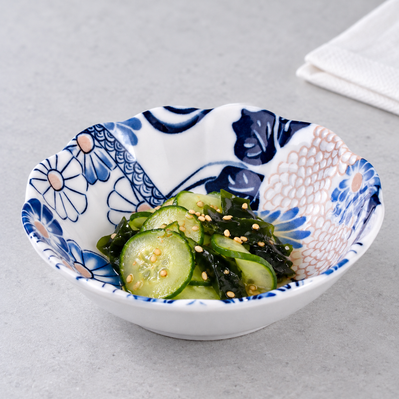 Shallow blue floral ceramic bowl with sliced cucumber, greens and scattered seeds on a light surface.