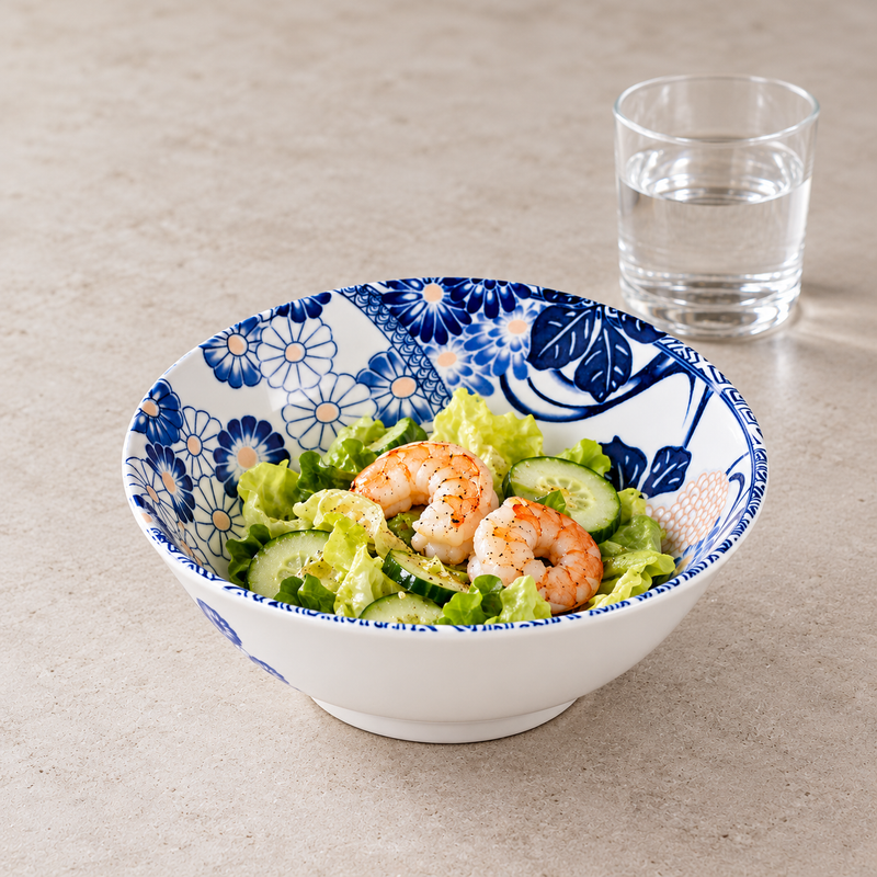 Blue floral ceramic bowl holding shrimp, lettuce and sliced cucumber, with a clear drinking glass behind it.
