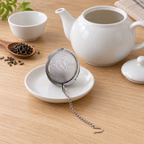 Closed V320 2.5-inch mesh tea ball and full chain on a separate dish beside a roomy open teapot and loose tea