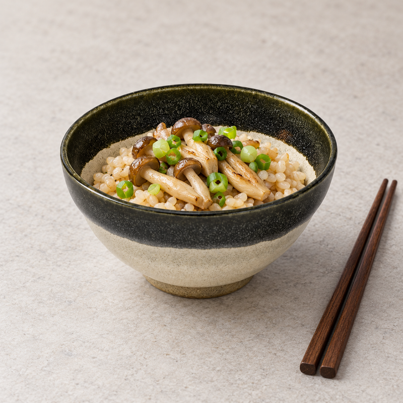 Cream ceramic bowl with a dark speckled rim holding rice, mushrooms and green additions beside brown chopsticks.