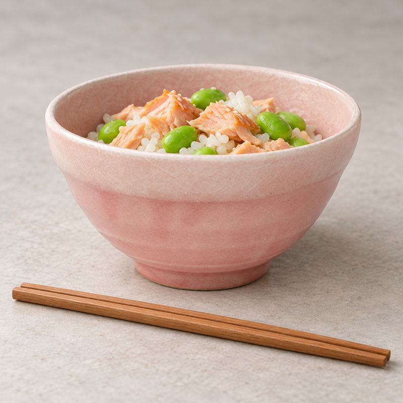 Pink and cream bowl holding rice with salmon and edamame, with chopsticks and other serving pieces nearby.
