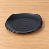 Black rounded-square melamine plate with subtle ripple detail on a tabletop