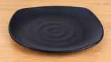 Black rounded-square melamine plate with subtle ripple detail on a tabletop