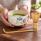 K537S-WR blue-floral matcha bowl set with bamboo whisk and scoop in a hand-held serving setting. Tea and surrounding scene accessories are illustrative, not included.