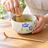 K537S-WR blue-floral matcha bowl set with bamboo whisk and scoop in a matcha preparation setting. Tea and surrounding scene accessories are illustrative, not included.