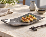MK212W-WB curved dark porcelain plate with three small gyoza, white brush motif exposed, beside sauce and chopsticks on a limestone table