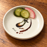 305-E Eggplant-pattern melamine sauce dish — Two cucumber pickle slices and one radish pickle slice at upper edge, leaving central motif mostly visible, warm wood tabletop